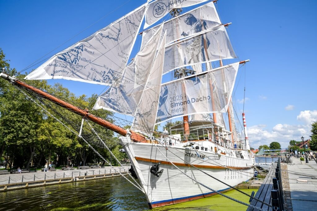A tall ship with white sails docked by a canal.